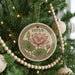 A round ornament with a pink lotus flower and green leaves is hanging on a Christmas tree, surrounded by ornaments and lights.