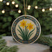 A decorative ceramic ornament featuring a floral design with yellow daffodils and green leaves, hanging from a string against a blurred background of greenery and twinkling lights.
