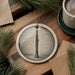 A ceramic ornament featuring a black and white illustration of a clarinet is displayed on a wooden surface, accompanied by a cup of coffee and some pine branches.