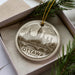 A circular ornament with a drawing of a city skyline, specifically Ottawa, Canada, is displayed in a box with pine branches.