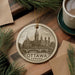 A decorative ornament featuring a cityscape of Ottawa, Canada, with a bridge and trees, is displayed on a wooden surface alongside a cup of coffee and some papers.