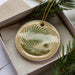 A decorative ceramic ornament with a palm tree design is displayed in a box, surrounded by pine branches.