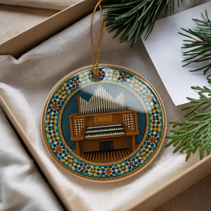 A decorative ornament featuring an organ, with a colorful border and a string hanging from it. The ornament is placed on a white surface, with some greenery visible in the background.