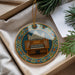 A decorative ornament featuring an organ, with a colorful border and a string hanging from it. The ornament is placed on a white surface, with some greenery visible in the background.