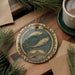 A decorative ornament featuring a zodiac symbol with two fish, placed on a wooden surface next to a cup of coffee and pine branches.