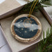 A decorative ornament with a starry night sky design and the word "Pleiades" on it, placed in a box with pine branches.