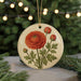 A round ornament featuring a red rose with green leaves, hanging from a string against a blurred background of a Christmas tree with lights.
