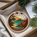 A decorative ornament with the text "SEYCHELLES" and "SEYCHELLES BEER" on it, hanging from a string. The ornament is placed on a white surface, with some greenery visible in the background.