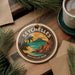 A round ornament with the text "SEYCHELLES" and "SEYCHELLES BEER" is displayed on a wooden surface, surrounded by pine branches and a cup of coffee.