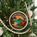 A round ornament on a Christmas tree, featuring a tropical beach scene with palm trees and a mountain in the background.