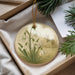 A decorative ornament featuring a floral design with snowdrops and a church in the background, placed in a box with pine branches.