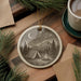 a decorative ceramic ornament depicting a camping scene with a tent, campfire, and mountains in the background, hanging on a wooden surface.