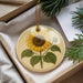 A round ornament with a sunflower design is placed in a box, surrounded by pine branches and greenery.