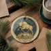 A decorative ornament featuring a bull, which is a zodiac animal, is displayed on a wooden surface alongside a cup of coffee and some pine branches.