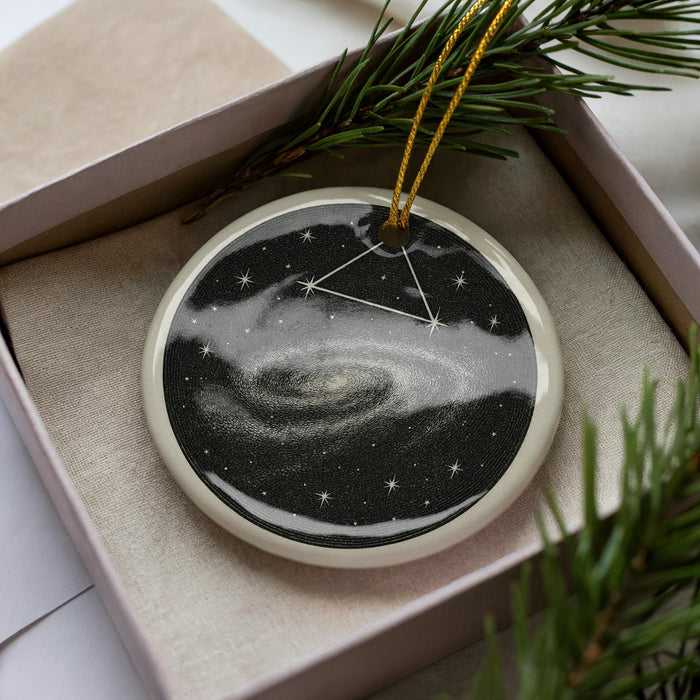 A circular ornament with a starry night sky design, hanging from a string and placed in a box with pine branches.