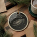 a circular ornament with a starry galaxy design, hanging on a wooden surface. The ornament is placed next to a cup of coffee and some pine branches.
