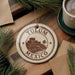 A circular ornament with the text "TULUM MEXICO" printed on it, surrounded by pine branches and a cup of coffee on a wooden surface.