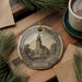 A decorative ornament featuring a church with a tower, surrounded by pine branches and a cup of coffee on a wooden surface.