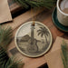 A decorative ornament featuring a guitar and a tropical landscape, placed on a wooden surface alongside a cup of coffee and some pine branches.