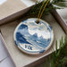 A decorative ornament in a box, featuring a blue and white design depicting a coastal landscape with mountains and waves.