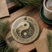 a decorative ornament with a yin and yang symbol, surrounded by pine branches and a cup of coffee on a wooden surface.