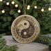 A decorative ornament featuring a yin and yang symbol, with a temple and mountain landscape in the center, is displayed on a table with pine branches and twinkling lights in the background.