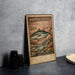 A metal stamp depicting a fish, likely Amberjack, with the text "Amberjack (GREATER)" and "20€ SERIOLA DUMERILI" printed on it, placed on a table next to some black camera lenses.