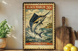 A vintage-style metal stamp depicting a black marlin fish, with the text "BLACK MARLIN 20¢" and "ISTIOPHORUS INDIA" printed on it. The stamp is displayed on a shelf next to a wooden cutting board and a potted plant.