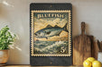 A framed vintage postage stamp featuring a bluefish is displayed on a shelf next to a wooden cutting board, a potted plant, and some lemons.