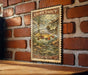 A framed print of a brown trout is hanging on a brick wall, depicting a fish swimming in a river.