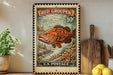 A vintage-style stamp depicting a red grouper fish, with the text "RED GROUPER 10¢" and "U.S. POSTAGE" printed on it. The stamp is displayed on a shelf next to a wooden cutting board and a potted plant.