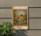 A vintage-style postage stamp featuring a rainbow trout, mounted on a wall next to a plant.