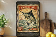 A vintage-style postage stamp featuring a swordfish, with the text "SWORDFISH AIR MAIL" and a price of 5 cents. The stamp is displayed on a wooden surface, with a potted plant, a cutting board, and some lemons nearby.