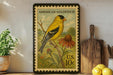 A framed American Goldfinch postage stamp is displayed on a shelf, with a potted plant, wooden cutting board, and lemons nearby.