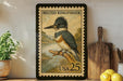 A framed postage stamp depicting a belted kingfisher, with the text "BELTED KINGFISHER" and "USA 25" printed on it. The stamp is placed on a shelf next to a potted plant, a wooden cutting board, and some lemons.