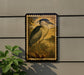 a framed painting of a heron standing on a grassy area, with a plant to the left of the frame.