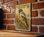 A vintage-style postage stamp featuring a colorful illustration of a bird perched on a branch, mounted on a brick wall.
