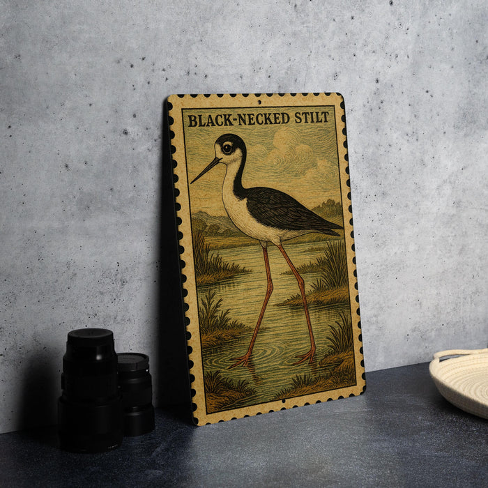 a metal sign with a black and white illustration of a bird, specifically a Black-Necked Stilt, standing on a grassy area near a body of water.