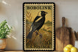 A vintage-style postage stamp featuring a black and white bird perched on a branch, with the text "BOBOLINK 10c U.S. POSTAGE" printed on it. The stamp is displayed on a shelf next to a wooden cutting board and a potted plant.