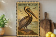 a framed illustration of a pelican, a large bird with a long beak, standing on a rock near the ocean. The pelican is depicted in a vintage style, with a brown and white color scheme. The framed image is placed on a shelf, surrounded by various kitchen utensils and items, such as a cutting board, a knife, and a bowl.