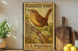 A vintage-style postage stamp featuring a brown bird perched on a branch, with the text "CAROLINA WREN" and "U.S. POSTAGE" printed on it. The stamp is displayed on a shelf next to a wooden cutting board and a potted plant.