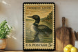A framed postage stamp depicting a loon is displayed on a shelf, surrounded by kitchen utensils and a potted plant.