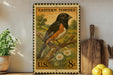 A vintage-style postage stamp featuring a colorful illustration of a bird perched on a branch, with a field and flowers in the background. The stamp is displayed on a shelf next to a wooden cutting board and a potted plant.