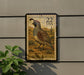 A postage stamp featuring a quail bird, with the number 22 and the text "USA" printed on it, mounted on a wall next to a plant.
