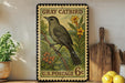 A vintage postage stamp featuring a gray catbird perched on a branch with flowers, placed on a wooden surface next to a wooden cutting board and a potted plant.