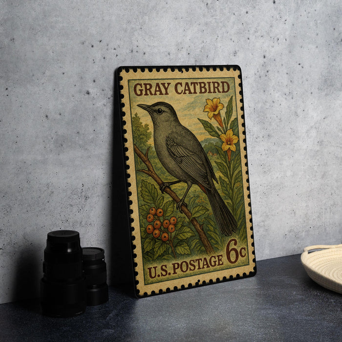 A vintage postage stamp featuring a gray catbird perched on a branch with flowers, placed on a countertop next to some black camera lenses.