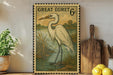 A framed postage stamp depicting a white heron standing in a pond, with a wooden paddle and a potted plant nearby.
