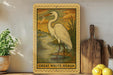 a framed illustration of a great white heron standing in a natural setting, with a wooden cutting board and a potted plant nearby.