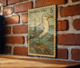 A framed postage stamp depicting a seagull is hanging on a brick wall.