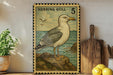 a framed illustration of a seagull, a type of bird, on a rock near the ocean. The seagull is depicted in a vintage style, with a postage stamp-like border. The seagull is standing on a rock, and there are some potted plants and a wooden cutting board visible in the background.